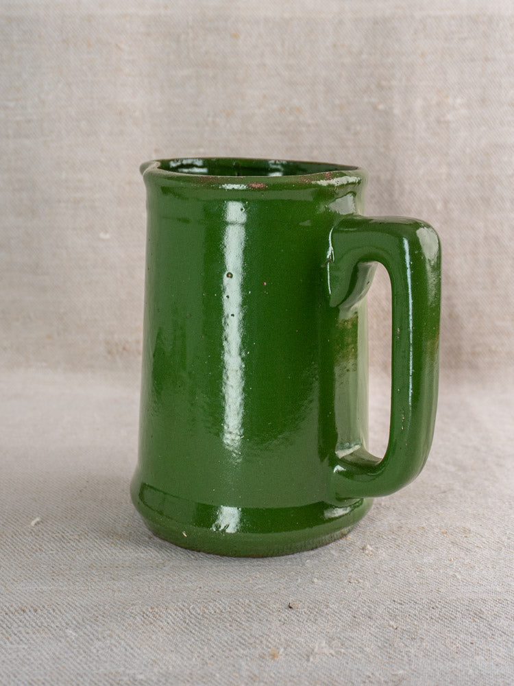 Antique French emerald green glazed earthenware pitcher with simple, flared shape on neutral fabric background.