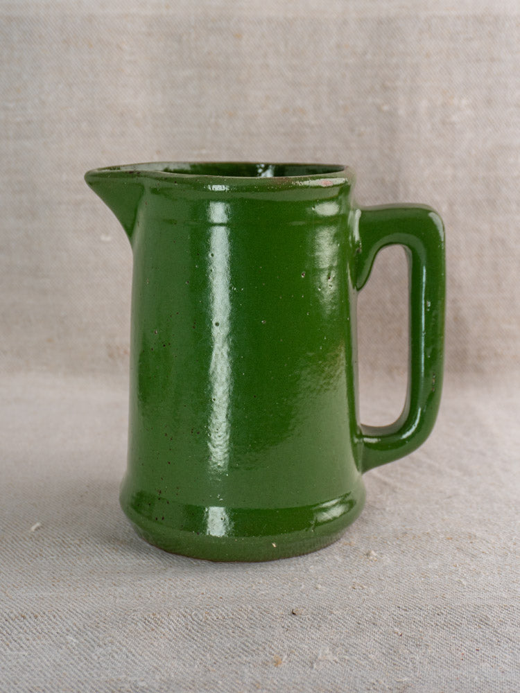 Antique French emerald green glazed earthenware pitcher with handle and spout on neutral textured fabric.