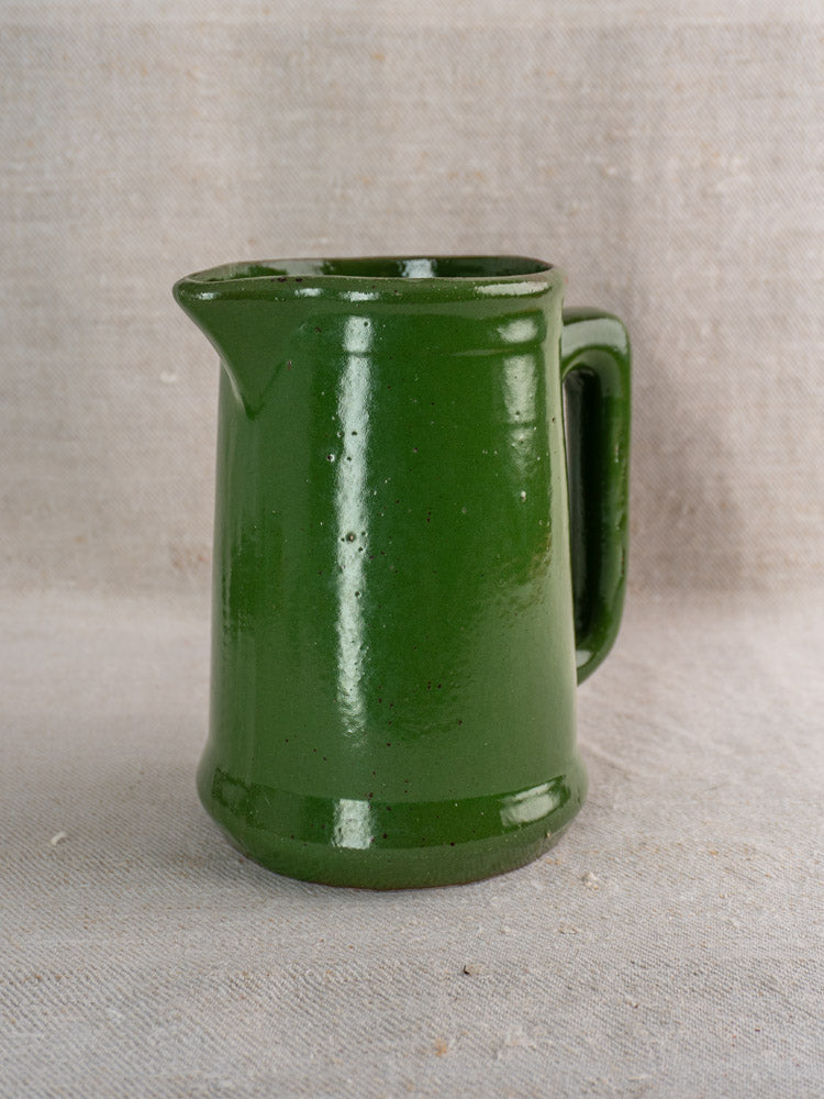 Antique French earthenware pitcher in deep emerald green glaze with handle and spout on beige fabric.
