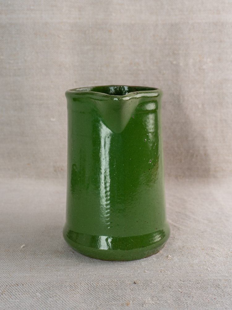 Small emerald green glazed antique French earthenware pitcher with spout on neutral textured fabric background.