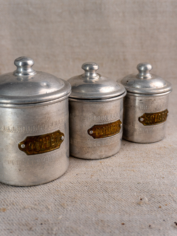 Antique French aluminum canister set of six with brass labels, timeworn patina, on textured beige fabric.