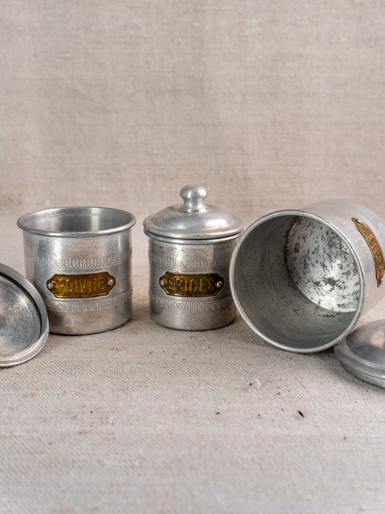 Antique French aluminum canister set with brass labels, Art Deco style, shown on textured beige fabric.