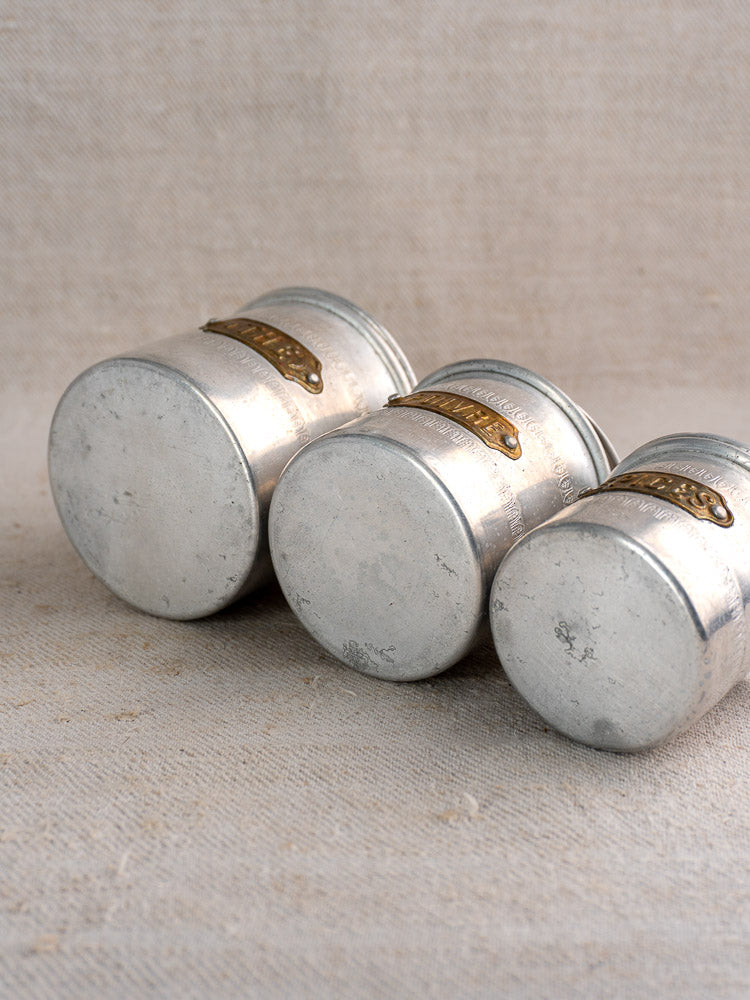Antique French aluminum canister set, 6 Art Deco pieces with brass labels on neutral textured fabric, bases facing up.
