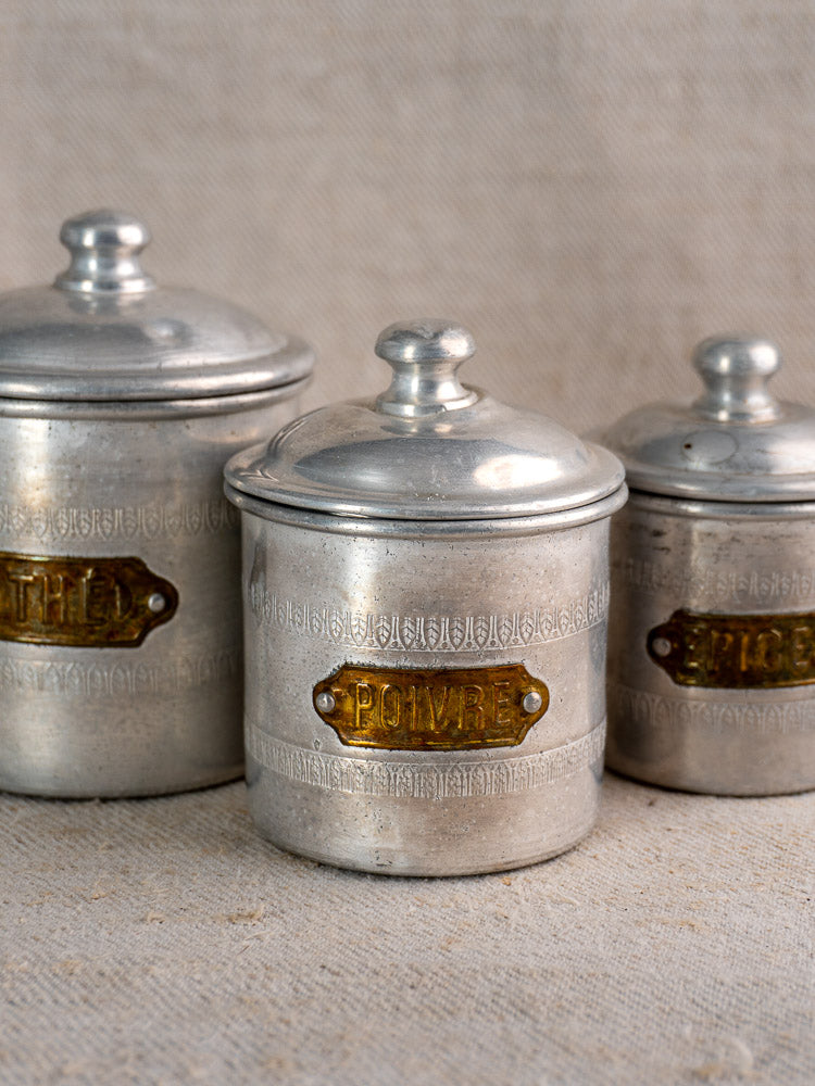 Antique French Art Deco aluminum canister set with brass labels on textured beige and fabric backdrop.