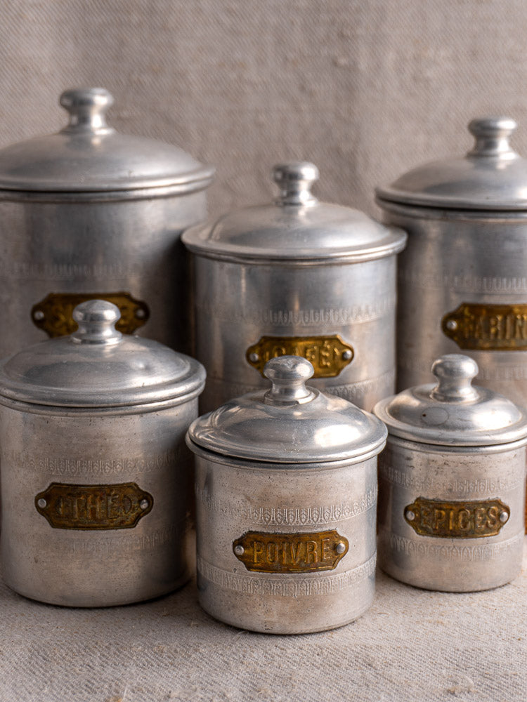 Antique French Art Deco aluminum canister set with brass labels, six lidded pieces on textured beige surface.