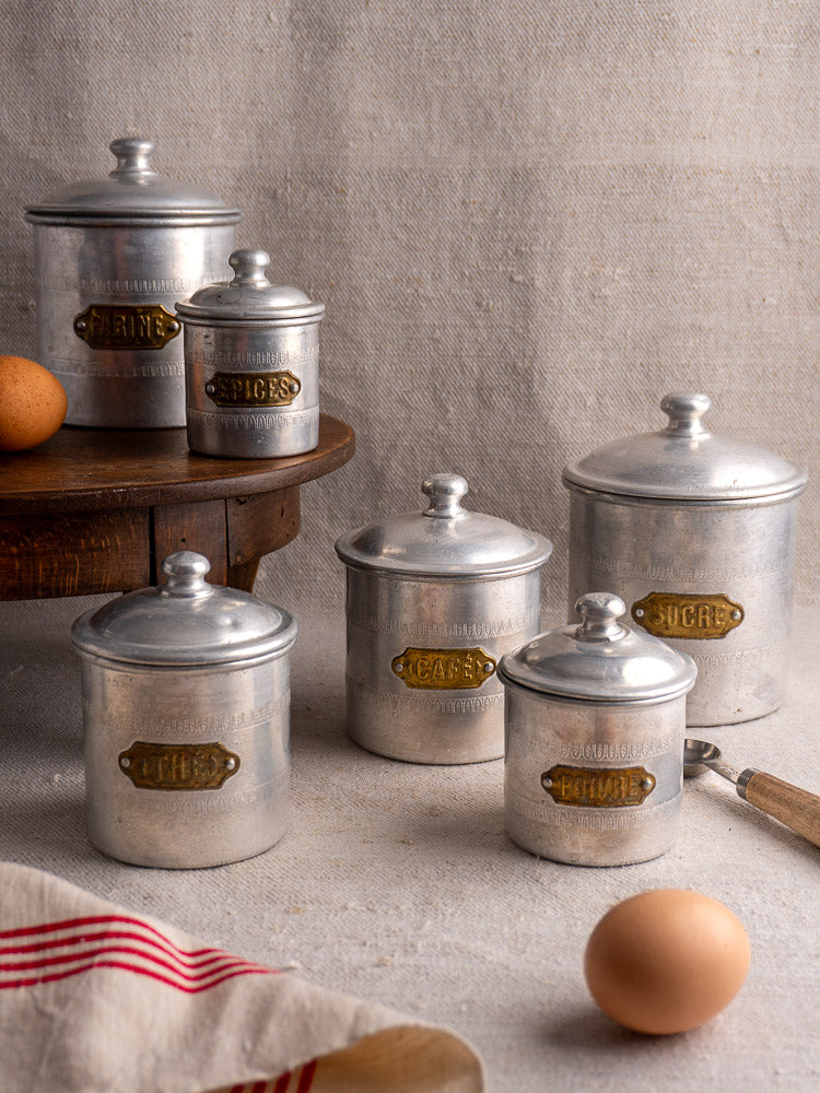 Antique French aluminum Art Deco canister set of six with brass labels on table, eggs, striped towel, wooden riser.