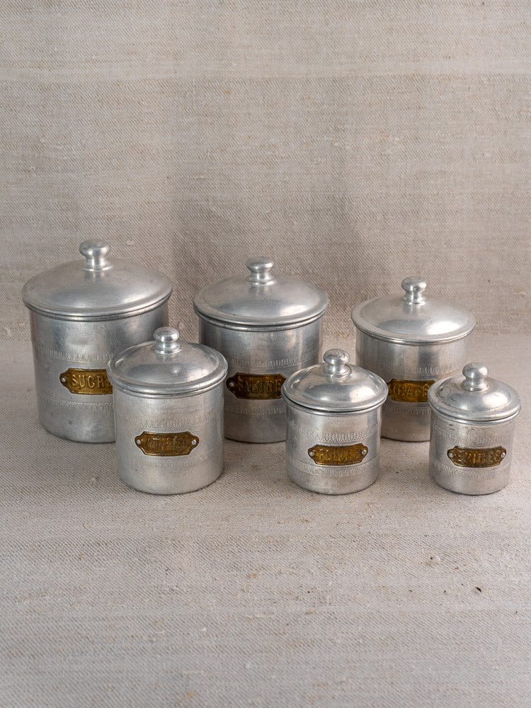 Antique French art deco aluminum canister set with brass labels, six pieces in two rows on neutral fabric.