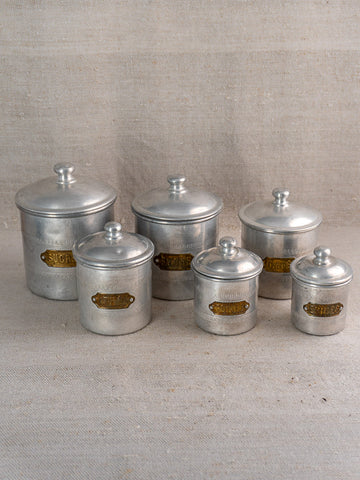 Antique French art deco aluminum canister set with brass labels, six pieces in two rows on neutral fabric.