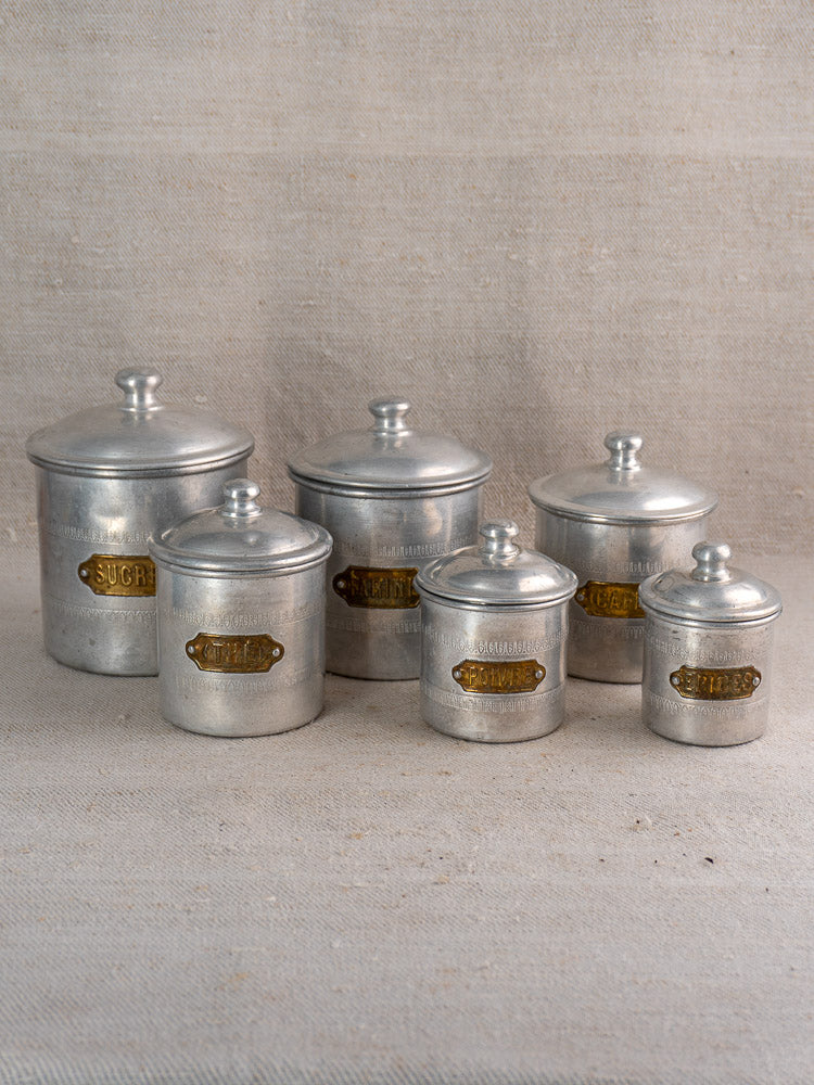 Antique French aluminum canister set, 6-piece art deco collection with brass labels on neutral fabric background.