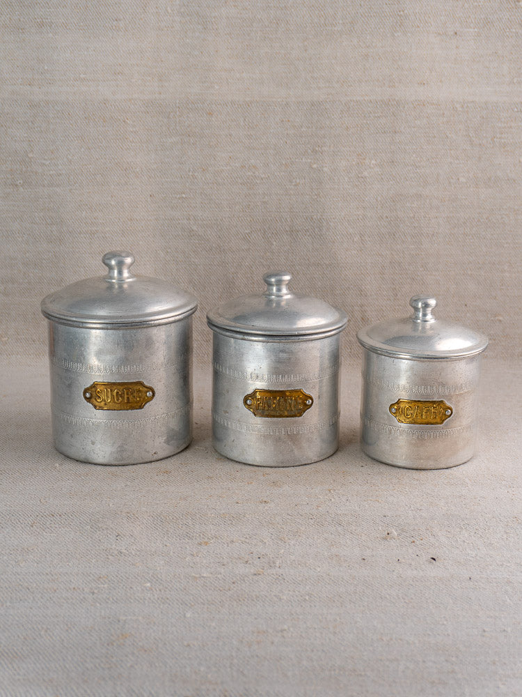 Antique French Art Deco 6-piece aluminum canister set with brass labels, nested on neutral textured background.