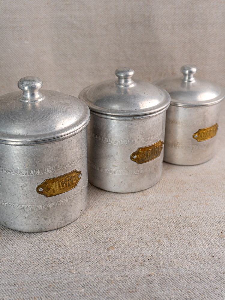 Antique French aluminum canister set, 6-piece Art Deco with brass labels and engraving on beige textured fabric.