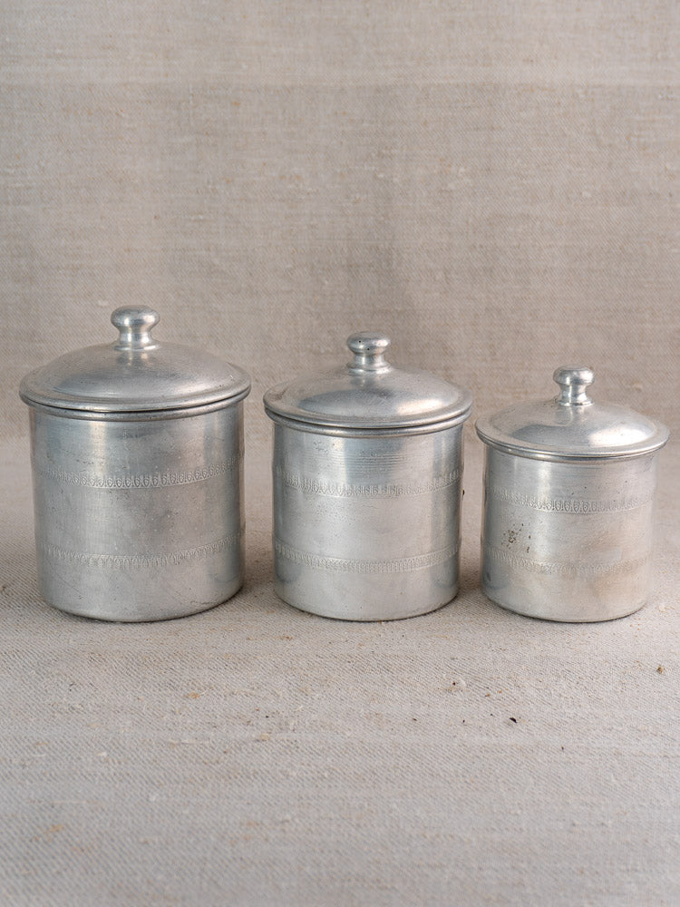 Antique French Art Deco aluminum canister set, 6 pieces with brass labels, on neutral fabric surface.