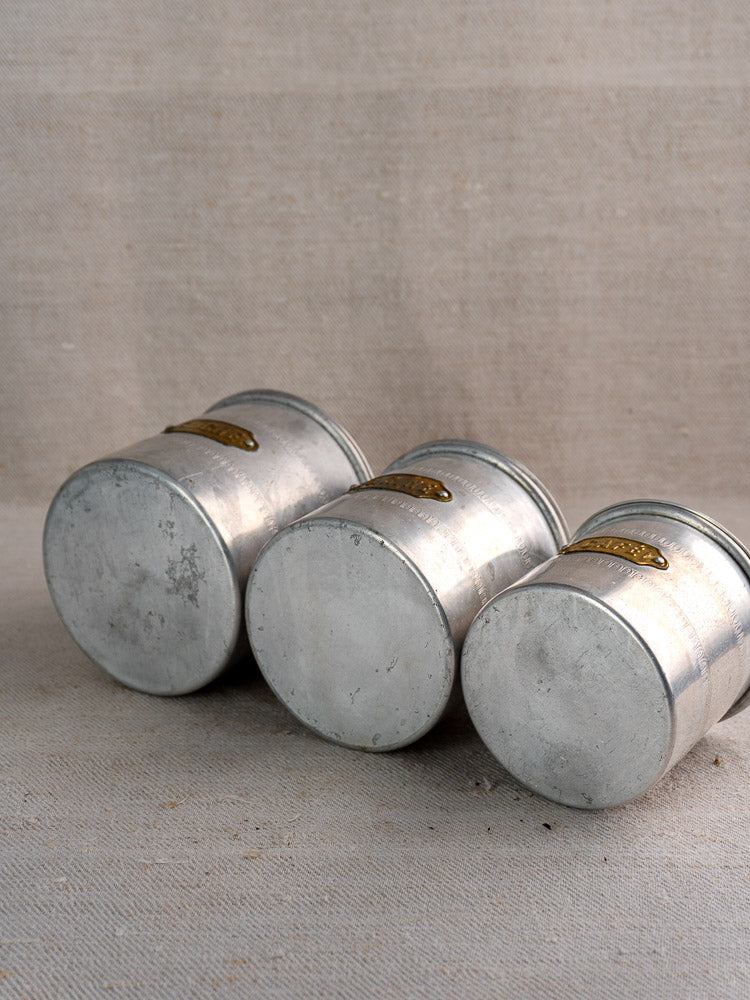 Antique French aluminum art deco canister set with brass labels on neutral textured fabric, highlighting vintage patina.