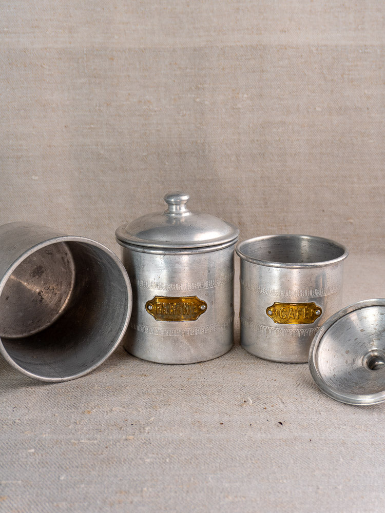 Art Deco antique French aluminum canister set of six with brass labels, arranged on neutral fabric against a beige backdrop.