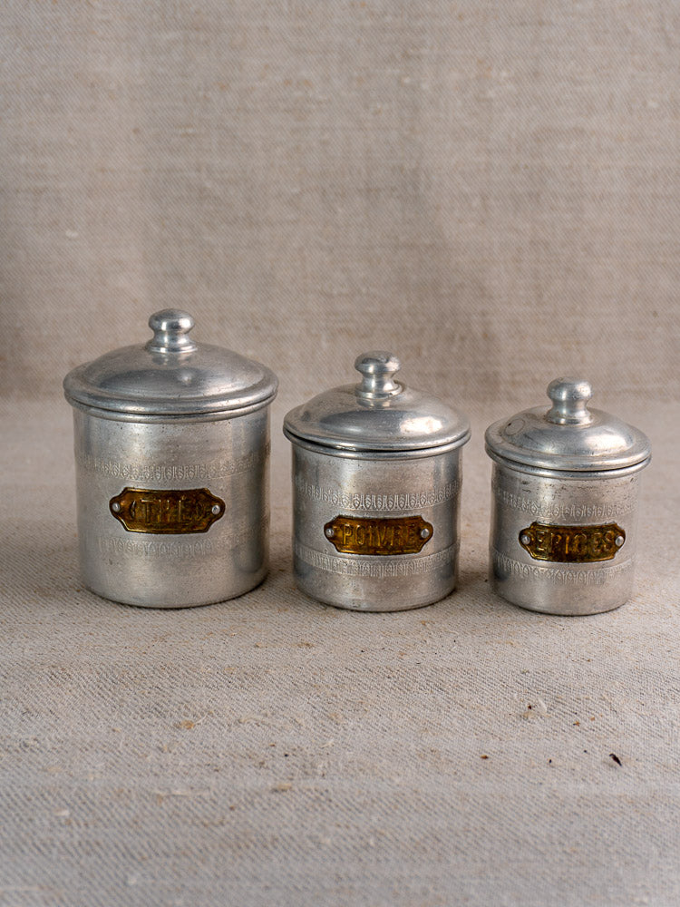 Antique French aluminum Art Deco canister set of six with brass labels, displayed on textured beige fabric.