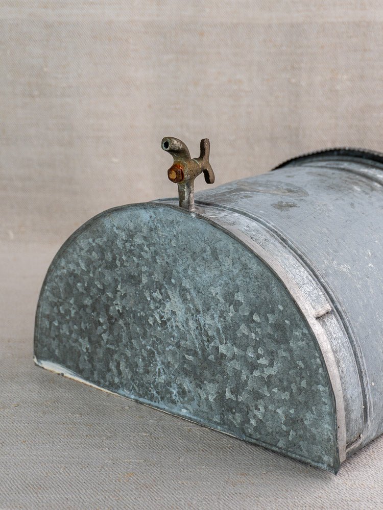Antique French zinc wall fountain with curved top and brass tap on beige textured surface.
