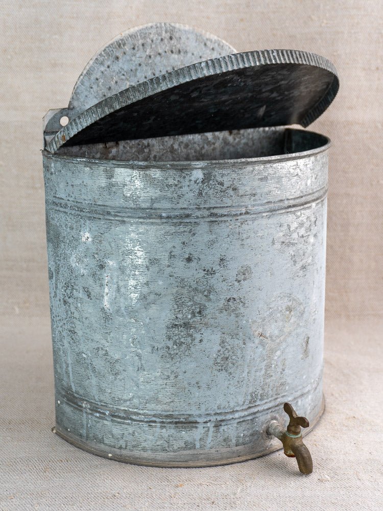 Antique French zinc wall fountain lavabo with round galvanized body, hinged lid, and brass tap on fabric.