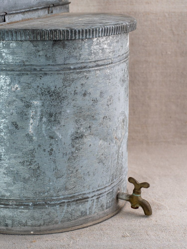 Antique French zinc wall fountain with brass tap, weathered finish, set against beige fabric.