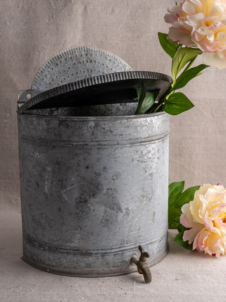 Antique French zinc wall fountain with brass tap, partially open lid, pink faux flowers, neutral fabric backdrop.