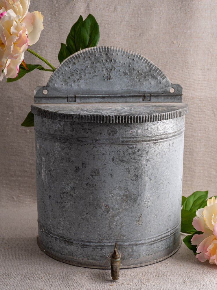 Antique French zinc wall fountain with ornate perforated lid and brass tap, accented by flowers.