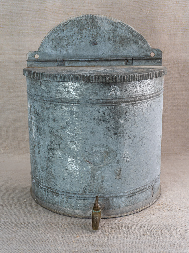 Antique French zinc wall fountain lavabo with ribbed semicircular back and brass tap, neutral fabric backdrop.