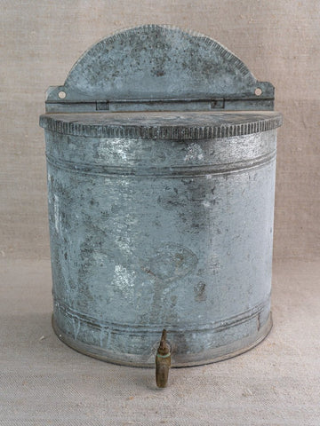 Antique French zinc wall fountain lavabo with ribbed semicircular back and brass tap, neutral fabric backdrop.