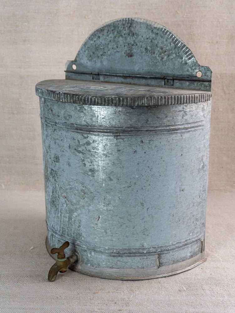 Antique French zinc wall fountain with brass tap and curved lid, early 20th century, on neutral fabric.