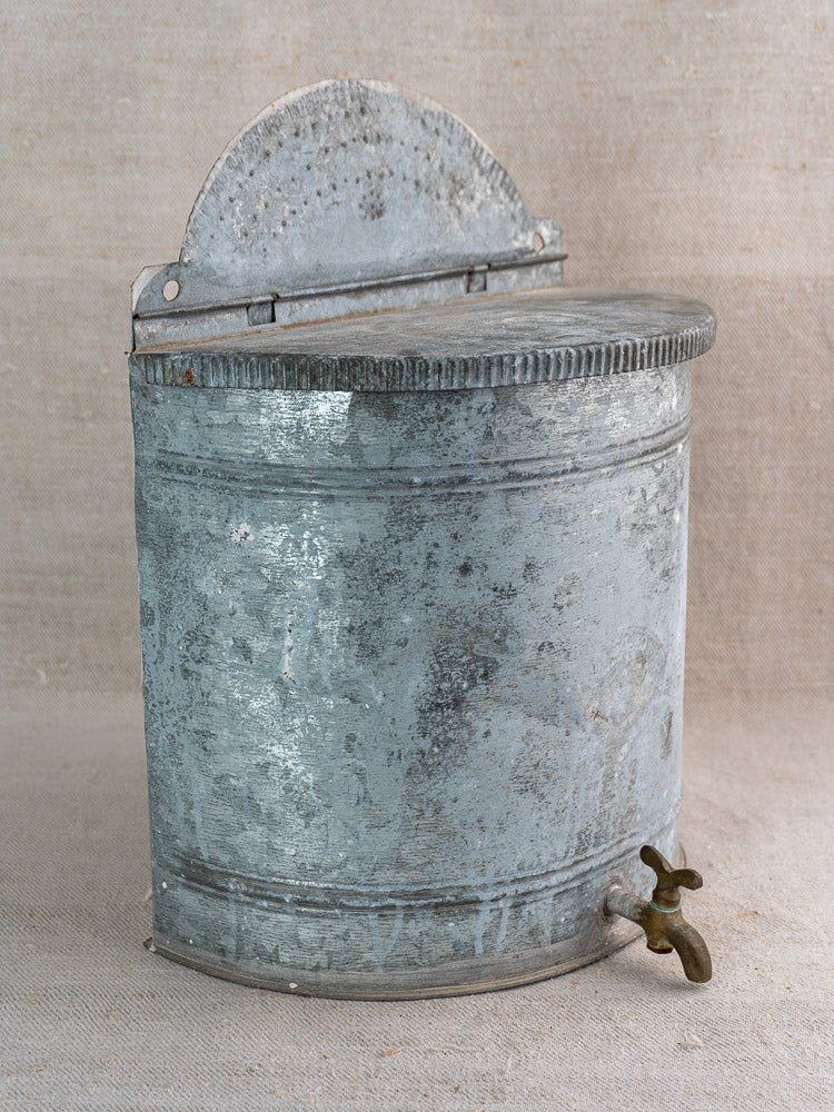 Antique French zinc wall fountain lavabo with curved lid and brass tap, against burlap background.