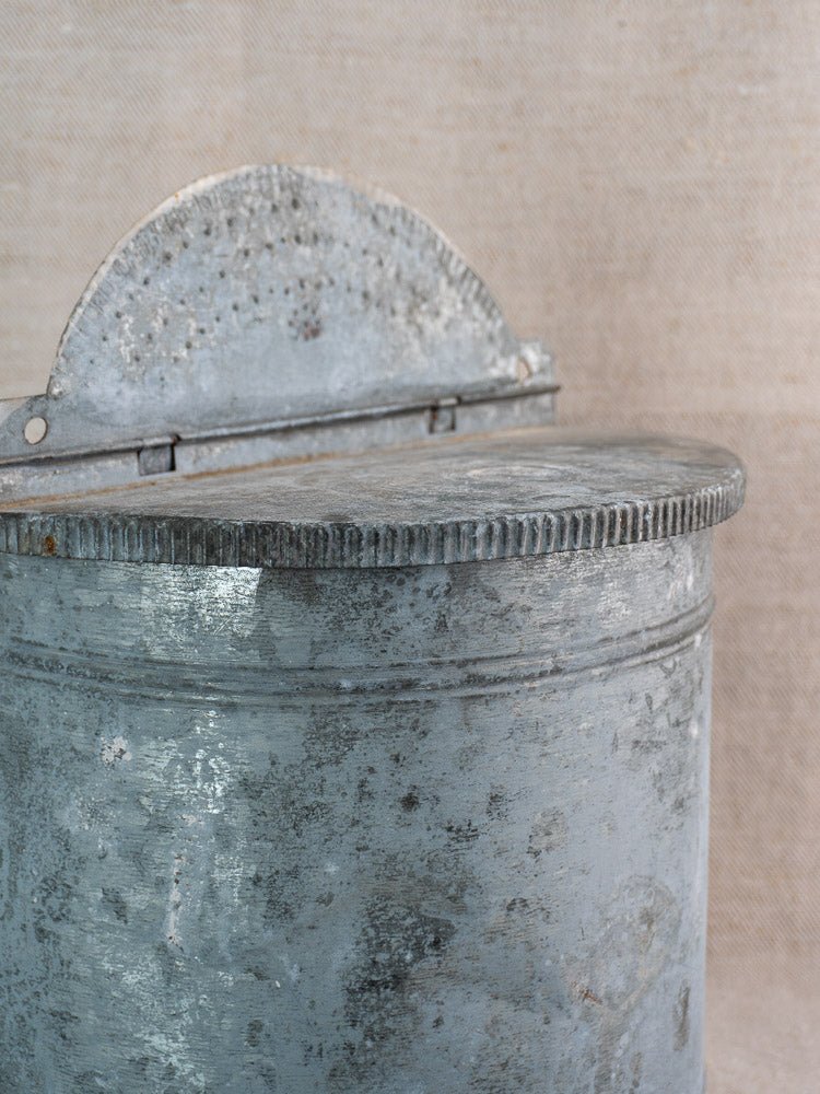 Antique French zinc wall fountain with brass tap, round weathered form, semi-circular lid on beige fabric.
