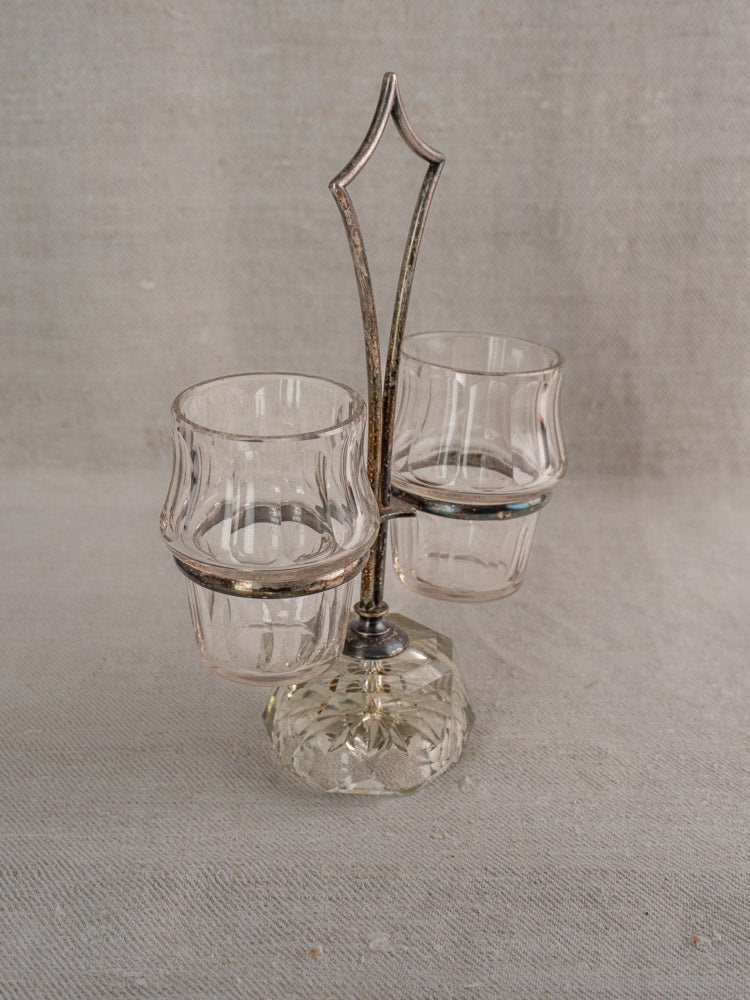 Antique French condiment server with two faceted cut glass cups in a decorative silver plate holder on light textured fabric.