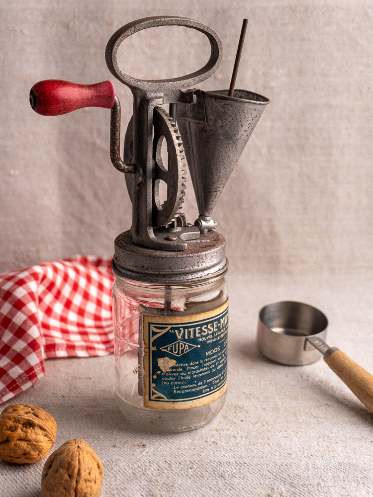 Antique French Vitesse Baratte mayonnaise maker with metal hand crank on glass jar, neutral fabric background.