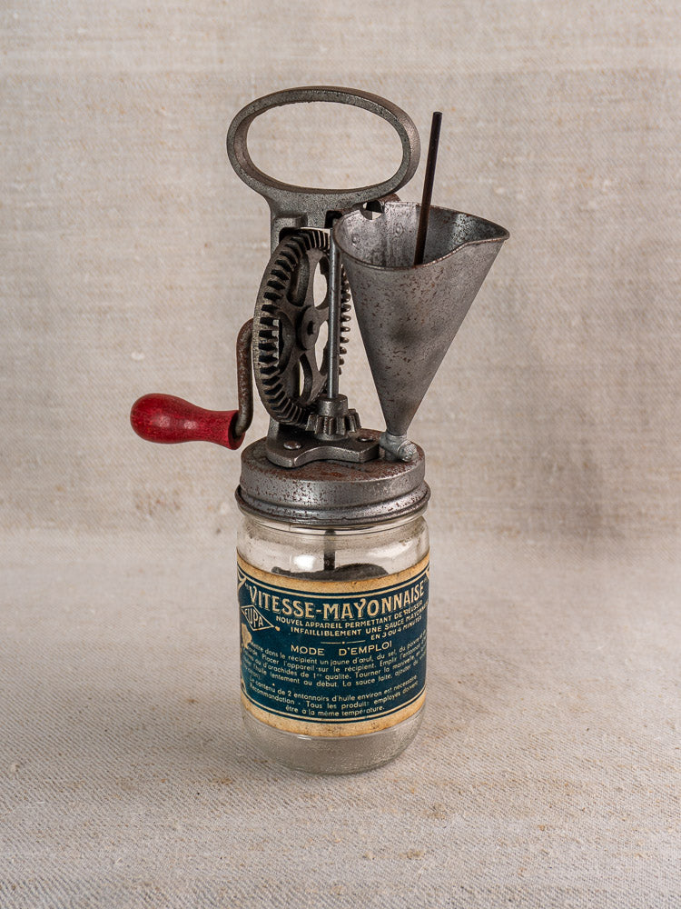 Antique French Vitesse Baratte mayonnaise maker with metal hand crank on glass jar, neutral fabric background.