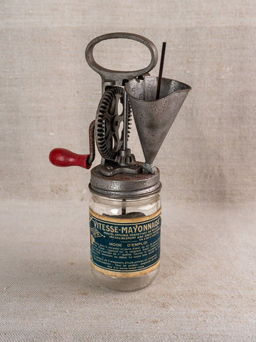 Antique French Vitesse Baratte mayonnaise maker with metal hand crank on glass jar, neutral fabric background.