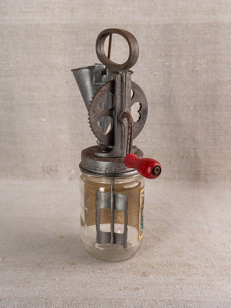 Antique French FUPA Vitesse Baratte mayonnaise maker with metal gears and red handle on glass jar, neutral fabric background.