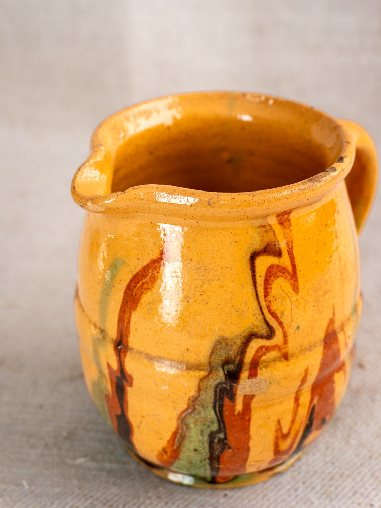 Antique French Jaspé pitcher with brown, green, and red marbling on yellow-orange clay, shown on a neutral textured surface.
