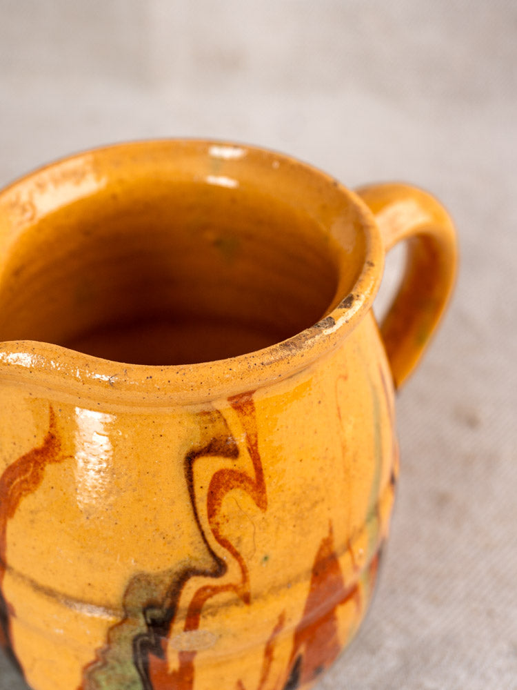 Antique French Jaspé pitcher with hand-thrown marbled slip glaze, orange, brown, green, handle; on neutral textured surface.