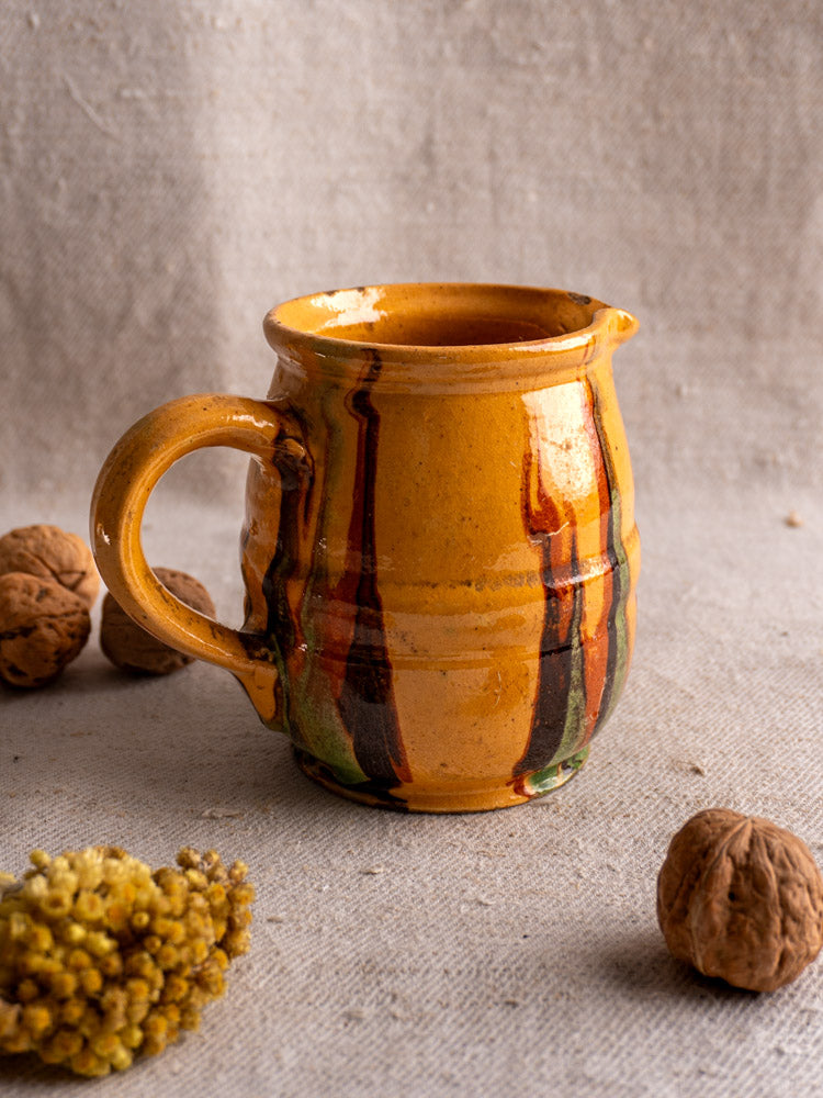 French antique Jaspé pitcher with earthy stripes and handle on beige cloth, walnuts and dried yellow flowers nearby.