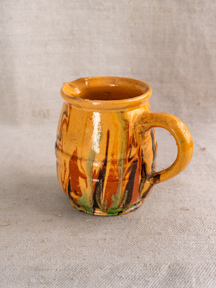 Antique French Jaspé pitcher with yellow-brown slip glaze and swirled brown, green, and orange on textured fabric surface.