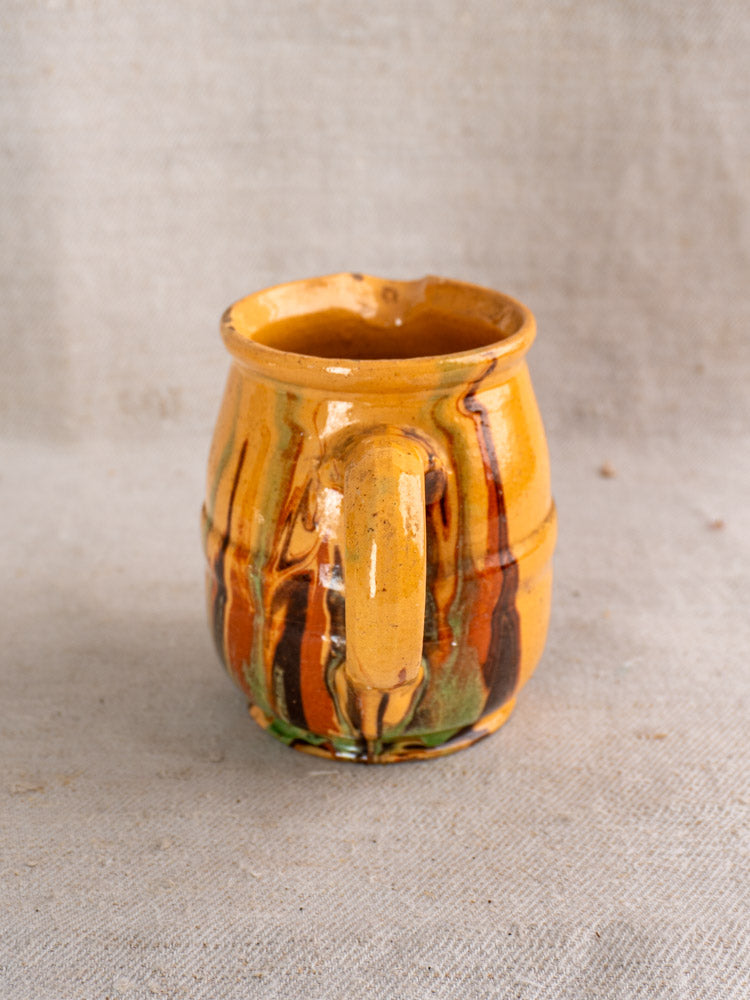Antique French Jaspé pitcher with yellow, brown, and green slip glaze, large handle, on textured neutral fabric.