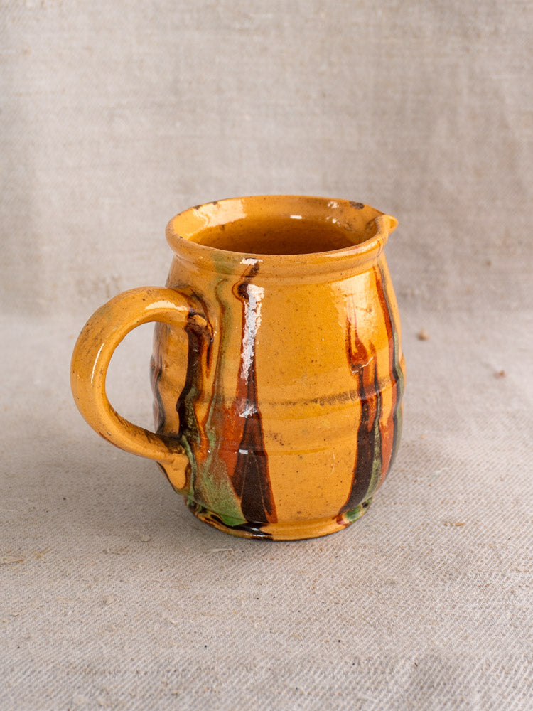 Antique French Jaspé pitcher, hand thrown, slip-glazed yellow and green-brown, late 19th century, on beige textured fabric.
