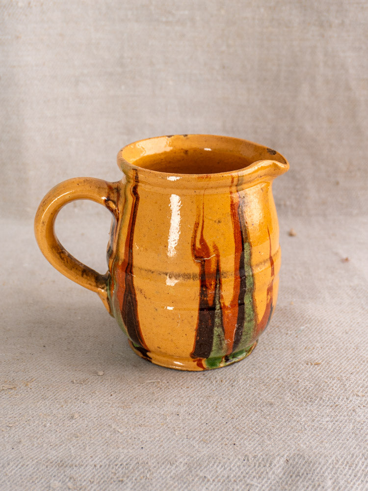 Late 19th-century French Jaspé pitcher with glossy yellow-orange glaze, brown and green streaks, on textured fabric.