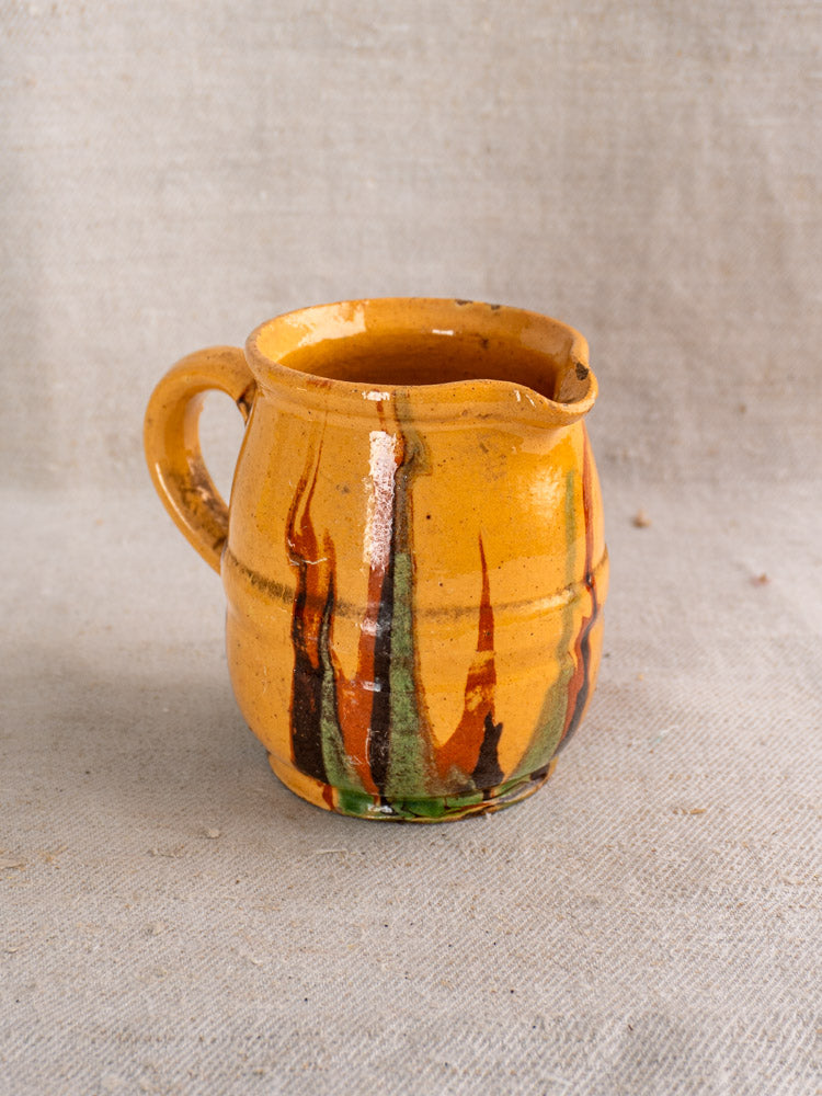 Antique French Jaspé ceramic pitcher with orange slip glaze and vibrant vertical streaks on beige fabric background.