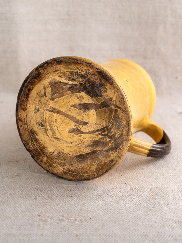 Antique French Pichet Jaune yellow glazed earthenware pitcher with worn base, small handle on beige fabric background.