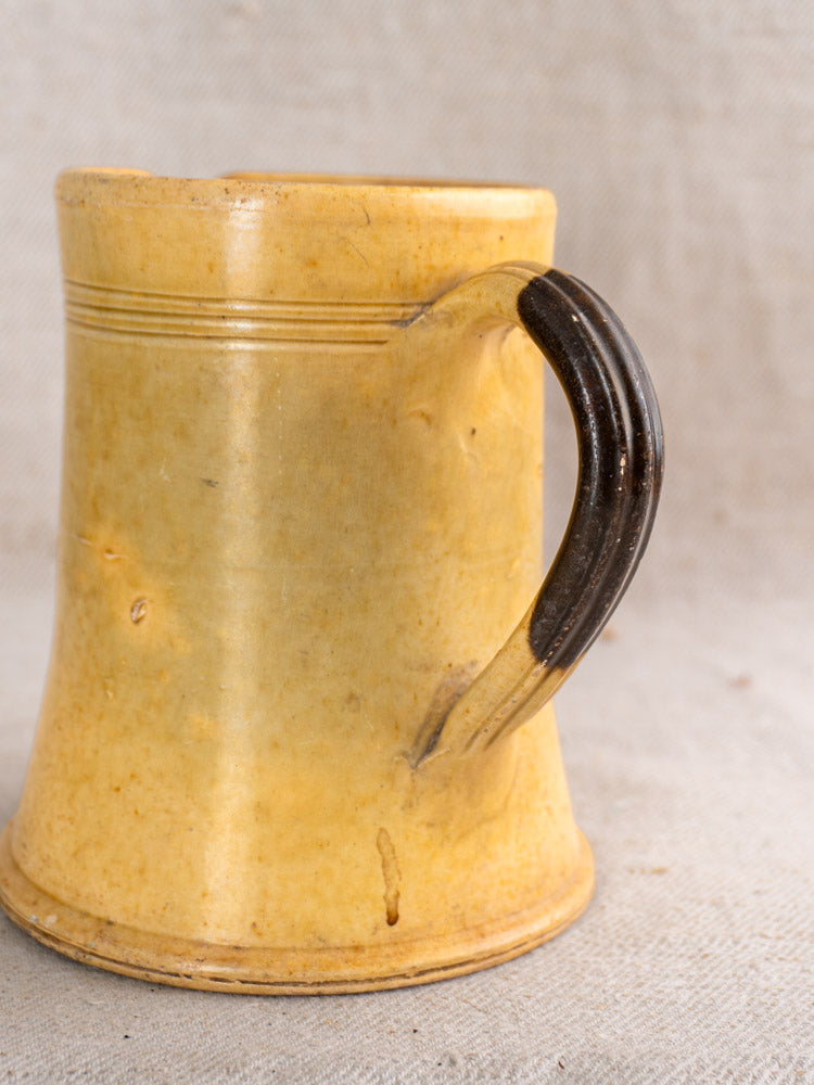 Antique French Pichet Jaune, yellow glazed earthenware mug with wide base and dark brown handle on beige textured surface.