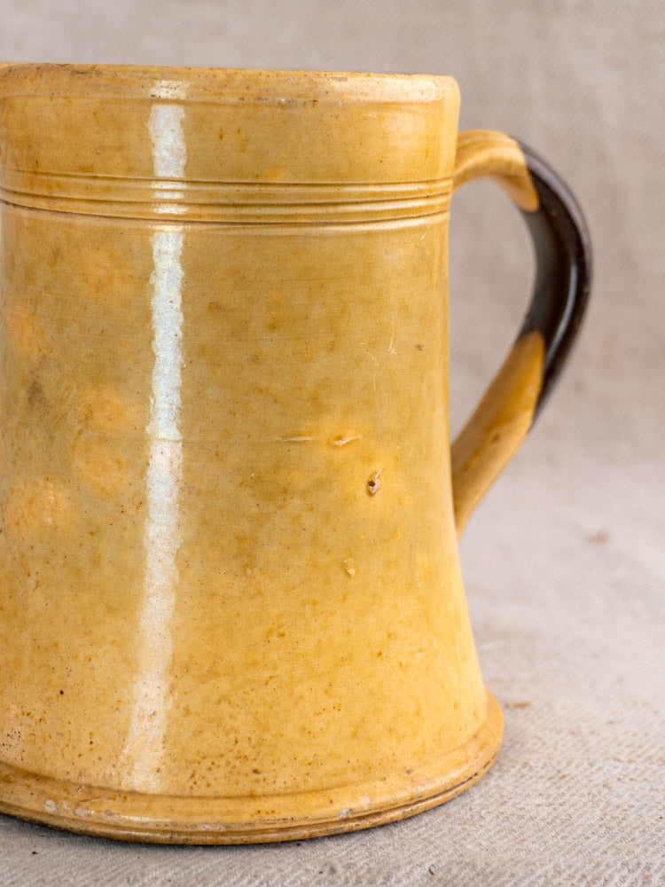 Antique French Pichet Jaune, yellow glazed earthenware mug with dark brown handle, rim grooves, on beige textured fabric.