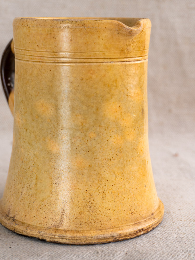 Antique French Pichet Jaune, yellow glazed earthenware mug with spout and handle, worn speckled finish on beige fabric.