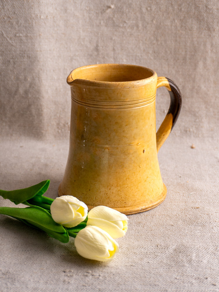 Antique French Pichet Jaune, yellow glazed earthenware pitcher, on beige fabric beside three white tulips.