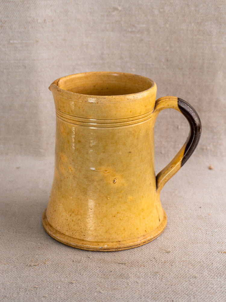 Antique French yellow glazed earthenware pichet with brown handle, flared base, on textured beige fabric.