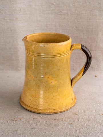 Antique French yellow glazed earthenware pichet with brown handle, flared base, on textured beige fabric.