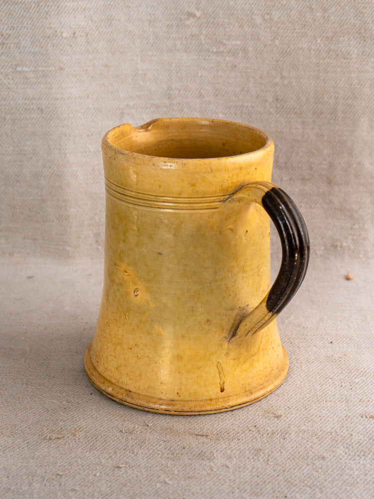 Antique French Pichet Jaune, yellow glazed earthenware mug with black handle and rim chip on beige textured fabric.